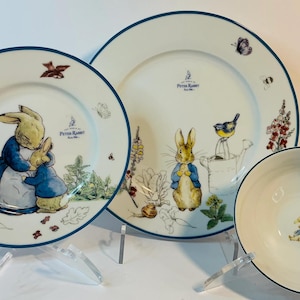 May include: A set of Peter Rabbit themed plates and a bowl. The plates and bowl are white with a blue trim. The plates feature illustrations of Peter Rabbit and other characters from the story. The bowl has a single Peter Rabbit illustration.