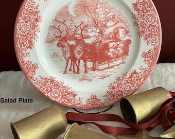 May include: A white ceramic salad plate with a red transferware pattern featuring Santa in a sleigh pulled by reindeer, a floral border, and a Royal Stafford Made in England stamp. Three gold-colored metal bells with red ribbons are positioned below.