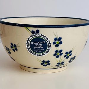 Puede incluir: Cuenco de cerámica polaca de color crema con borde azul oscuro y diseño floral. El cuenco presenta flores azules pintadas a mano con detalles verdes. Una etiqueta circular indica "BOLESLAWIEC POLISH POTTERY Hand Painted in Poland."