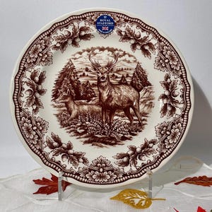 May include: A cream-colored ceramic plate featuring a brown transferware print of a stag and a doe in a forest setting. The rim is decorated with a repeating pattern of oak leaves and acorns. A blue Royal Stafford logo is printed at the top.
