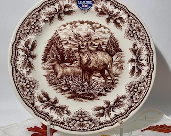 May include: A cream-colored ceramic plate featuring a brown transferware print of a stag and a doe in a forest setting. The rim is decorated with a repeating pattern of oak leaves and acorns. A blue Royal Stafford logo is printed at the top.