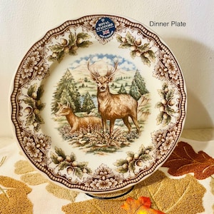 May include: A decorative dinner plate with a brown and cream floral border. The center features a scenic illustration of two deer in a forest setting. The plate has a scalloped edge and a Royal Stafford stamp.