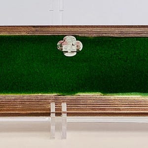Puede incluir: Una bandeja rectangular de madera con interior de fieltro verde oscuro. La bandeja tiene un marco de madera de color claro y un pequeño emblema decorativo plateado en el centro. La bandeja está soportada por soportes de plástico transparente.