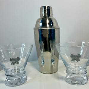 Puede incluir: Una coctelera de acero inoxidable con dos copas de martini transparentes y un adorno en forma de lazo. Las copas están sobre una superficie blanca. La coctelera está en el centro, con una copa a cada lado.