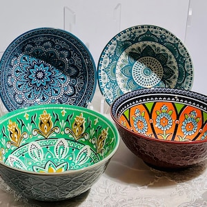 May include: A set of four ceramic bowls featuring intricate patterns. Two bowls are displayed upright in the back with blue and teal mandala designs. Two bowls are in the front, one with a green and yellow floral pattern and one with orange and blue flowers.