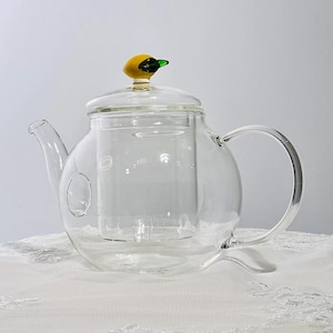 Puede incluir: Una tetera de vidrio transparente con un cuerpo redondeado y un asa curva. La tapa está rematada con un adorno en forma de pato amarillo y verde. La tetera tiene un infusor incorporado y un pico vertedor.