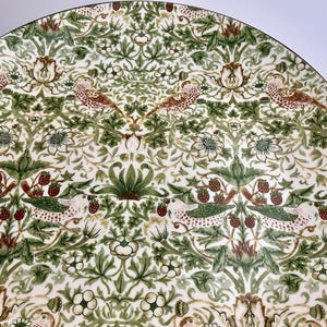 Op de afbeelding: Een decoratieve keramische plaat met een gedetailleerd patroon. Het ontwerp toont vogels, aardbeien en bloemelementen in groen, bruin en wit. De plaat heeft een gouden rand, wat een vleugje elegantie toevoegt.