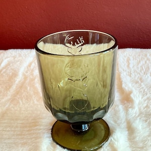 May include: Olive green glass goblet with a deer head design. The glass has a short stem and a wide, rounded bowl. The deer head is etched into the glass. The goblet is sitting on a white, fluffy surface.