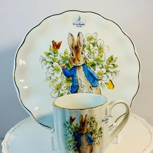 May include: A Peter Rabbit themed tea set. The white plate has a scalloped edge and gold trim, featuring a Peter Rabbit illustration. The mug also has gold trim and a Peter Rabbit illustration. The set is ideal for a tea party.