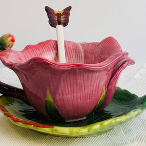 Può includere: Una tazza da tè in ceramica rosa a forma di fiore, con un piattino abbinato a forma di foglia. Un cucchiaino con un farfalla è appoggiato nella tazza. Il manico e il bocciolo sono verdi e marroni.