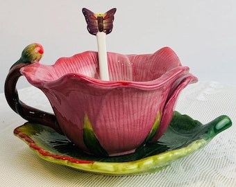 May include: A pink ceramic teacup shaped like a flower, with a matching leaf-shaped saucer. A butterfly-topped spoon sits in the cup. The handle and bud are green and brown.