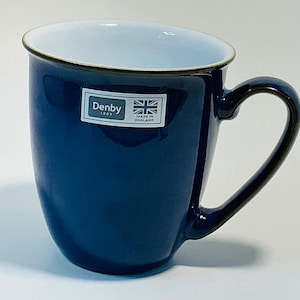 Pode incluir: Uma caneca de cerâmica azul marinho com interior branco e borda marrom escuro. A caneca tem uma alça curva e um rótulo que diz "Denby 1809 Made in England". Ideal para bebidas quentes.