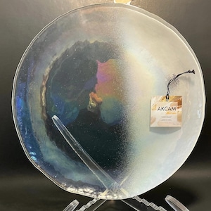 May include: Iridescent glass plate with a unique, organic shape. The plate has a shimmering, multi-colored surface with hints of blue, green, and gold. A tag with the text "AKCAM" is attached. The plate is displayed on a clear stand.