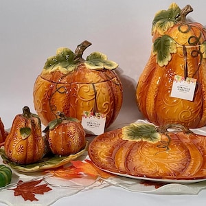 Puede incluir: Una colección de utensilios de cocina de cerámica con forma de calabaza que incluye una mantequera, salero y pimentero, un tarro grande, una bandeja y una jarra de leche. Cada pieza tiene un esmalte naranja y detalles de hojas verdes.