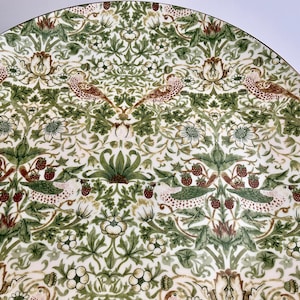 May include: A round ceramic plate featuring a repeating pattern of birds, strawberries, and foliage in shades of green, brown, and cream. The design includes small red strawberries and white flowers set against a dense background of leaves.