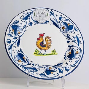 May include: White ceramic plate with a blue floral border and a central image of a rooster. The rooster is yellow, orange, and brown, standing on green grass. The plate has the text "MADE IN ITALY ITALIE" on it.