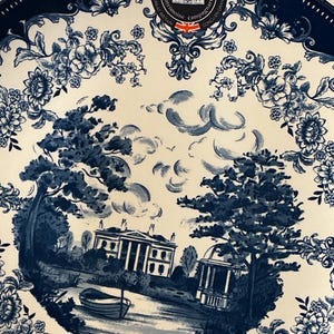 May include: A white and blue decorative plate with a scenic landscape design. The plate features a detailed illustration of a building, trees, and a body of water. The rim of the plate has a blue floral pattern. The text "Eaton Fine Dining" is visible.