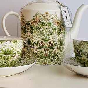 Op de afbeelding: Een wit keramisch theeservies met een theepot, twee kopjes en schotels. De set is versierd met een groen en crèmekleurig bloemmotief. De kopjes en de theepot hebben het label "MORRIS & CO" bevestigd.
