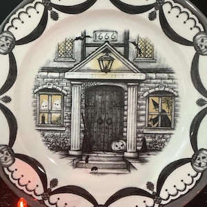 May include: A round ceramic plate featuring a black and white illustration of a house with the year 1666 above the door. The scene includes ghosts in a window, a witch in another, a pumpkin, and a border decorated with repeating pumpkin motifs.