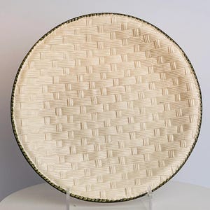 May include: A round, off-white plate with a woven basket weave pattern. The plate has a dark green trim around the edge. The plate is displayed on a clear stand.