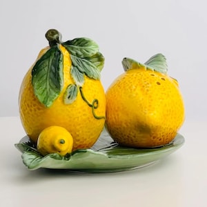 Puede incluir: Un juego de salero y pimentero de cerámica con forma de limón. El juego incluye dos saleros amarillos con hojas verdes y un limón pequeño, todo sobre un plato verde con forma de hoja. Los saleros tienen una superficie texturizada.
