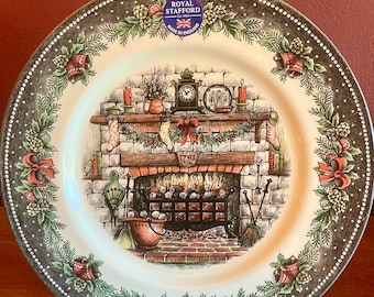 May include: A decorative plate with a Christmas theme. The plate features a detailed illustration of a fireplace with stockings, candles, and a clock. The border has a floral and bell design with the text "Christmas Evening".