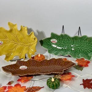 May include: Three ceramic leaf-shaped serving dishes in yellow, green, and brown. Each dish features a detailed embossed vein pattern and a glossy finish. A small white sticker with the text Hand Painted is attached to the surface of each dish.