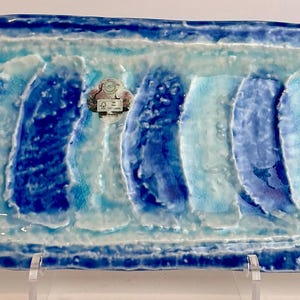 Puede incluir: Bandeja rectangular de cerámica en azul y blanco. La bandeja presenta una superficie texturizada con bandas alternas de azul claro y oscuro, y un borde elevado. La bandeja está soportada por un soporte transparente.