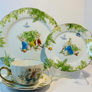May include: A Peter Rabbit themed tea set featuring a teacup, saucer, and two plates. The white porcelain pieces have a gold trim and illustrations of Peter Rabbit in a garden setting. Green foliage and floral patterns decorate the edges.