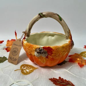 May include: A ceramic pumpkin-shaped basket with a cream-coloured interior and a textured exterior featuring orange and red autumn leaves and acorns. The handle resembles a gnarled branch. A brown paper tag reads Celebrate Fall.