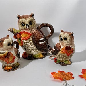 May include: A ceramic tea set featuring three owl-shaped pieces with brown, white, and tan feathers. The set includes a teapot, a creamer, and a sugar bowl, each decorated with orange and yellow autumn leaves and small brown acorns.