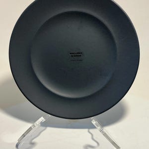 May include: A matte black dinner plate with a slightly raised rim. The plate is displayed on a clear acrylic stand. The text "every place is unique" and "made in Portugal" is printed on the bottom of the plate.