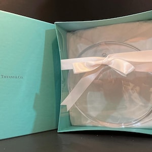 Op de afbeelding: Een heldere glazen plaat wordt gepresenteerd in een Tiffany & Co. doos. De plaat is verpakt in vloeipapier en vastgebonden met een wit satijnen lint. De doos is Tiffany blauw.