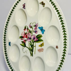 Op de afbeelding: Ovale, witte keramische serveerschaal met tien eivormige inkepingen. De schaal heeft een bloemenontwerp van roze en paarse lathyrus, vlinders en groene bladaccenten. De rand is versierd met een groen bladerpatroon.
