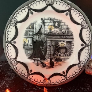 May include: A round white ceramic plate featuring a black and white illustration of a witch in a pointed hat standing before a fireplace with a cauldron. The year 1666 is inscribed above the hearth. The border is decorated with pumpkin motifs.
