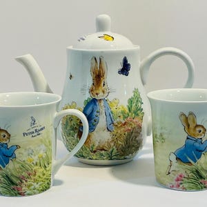 Può includere: Servizio da tè in ceramica bianca con illustrazioni di Peter Coniglio. Il set include una teiera con coperchio e due tazze abbinate. La teiera e le tazze presentano immagini di Peter Coniglio con una giacca blu, circondato da fiori.