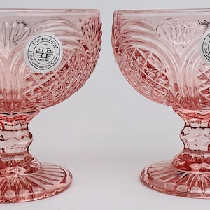 May include: Two pink crystal champagne coupe glasses with intricate diamond and floral patterns. Each glass has a round bowl, a stem with a decorative design, and a circular base. The glasses have a label that reads "Fitz and Floyd".