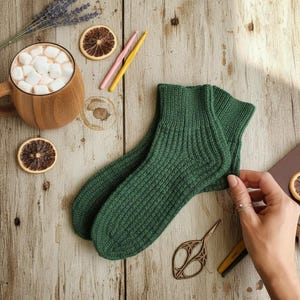 Peut inclure: Une paire de chaussettes vertes en tricot côtelé sur une surface en bois, avec une tasse de chocolat chaud garnie de guimauves, des tranches d'orange séchées et de la lavande. Une paire de ciseaux et un stylo sont également visibles.