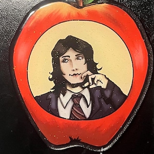 May include: An apple-shaped ornament featuring a portrait of a person in a suit and tie. The portrait is set against a yellow background within the apple's core. The apple is red with glitter accents.