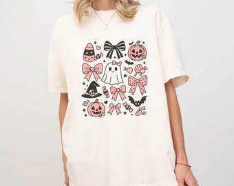 May include: Cream-colored t-shirt featuring a collection of Halloween-themed illustrations. The design includes pink bows, pumpkins, a ghost, a witch's hat, bats, and candy. The graphic is in black and pink, creating a festive and playful aesthetic.