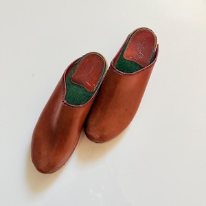Könnte beinhalten: Ein Paar braune Lederclogs mit runder Spitze. Der Innenraum hat grünen Stoff und eine braune Lederinnensohle. Die Clogs haben ein klassisches Slip-on-Design, geeignet für den Innen- oder Außenbereich. Die Clogs haben eine warme braune Farbe.