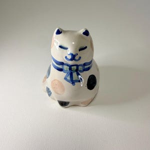 Puede incluir: Figura de gato de cerámica con base blanca, adornada con lunares azules y rosas. El gato lleva una pajarita azul alrededor del cuello. Las orejas y los ojos del gato también están pintados de azul. Es un objeto decorativo.