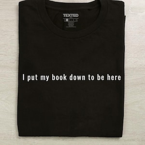 Könnte beinhalten: Schwarzes T-Shirt mit dem weißen Text "I put my book down to be here". Das T-Shirt hat einen Rundhalsausschnitt und ein Etikett oben. Das T-Shirt ist ordentlich gefaltet.