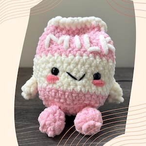 May include: A handmade crochet milk carton plushie in pink and white. The plushie has a smiling face with black eyes and pink cheeks. The word "MILK" is stitched across the front. The plushie has small arms and legs.