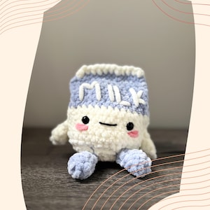 May include: A handmade crochet milk carton plush toy. The toy is white and light blue with the word "MILK" embroidered on the front. It has black eyes, pink cheeks, and small blue feet and arms. The background is a neutral color.