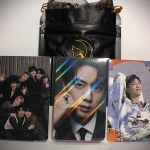 May include: Three rectangular photo cards featuring members of the K-pop group BTS, displayed in clear protective sleeves in front of a black pouch with a gold wolf and moon wax seal. The cards show a group portrait, a portrait of Jin, and RM.