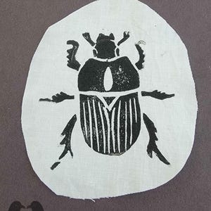 May include: A black scarab beetle design printed on a white, oval-shaped fabric patch. The beetle is detailed with vertical lines on its back and is set against a dark gray background. The patch is likely for decorative purposes.
