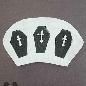 May include: A white fabric patch featuring three black coffins with white crosses and the number 4. The patch has a dark gray background and a stylized bird logo in the lower left corner. The design is likely for a goth or Halloween theme.
