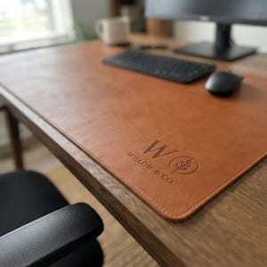 May include: A rectangular tan desk mat with rounded corners and a smooth surface. The mat features a debossed logo with the letter W, the text WILLOW & CO., and a circular icon containing a small leaf sprig, positioned on the bottom right corner.