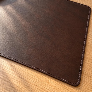 May include: A rectangular dark brown placemat with a textured surface and rounded corners. The edges of the placemat are finished with a decorative stitched border in a matching dark brown thread, creating a clean and uniform appearance.